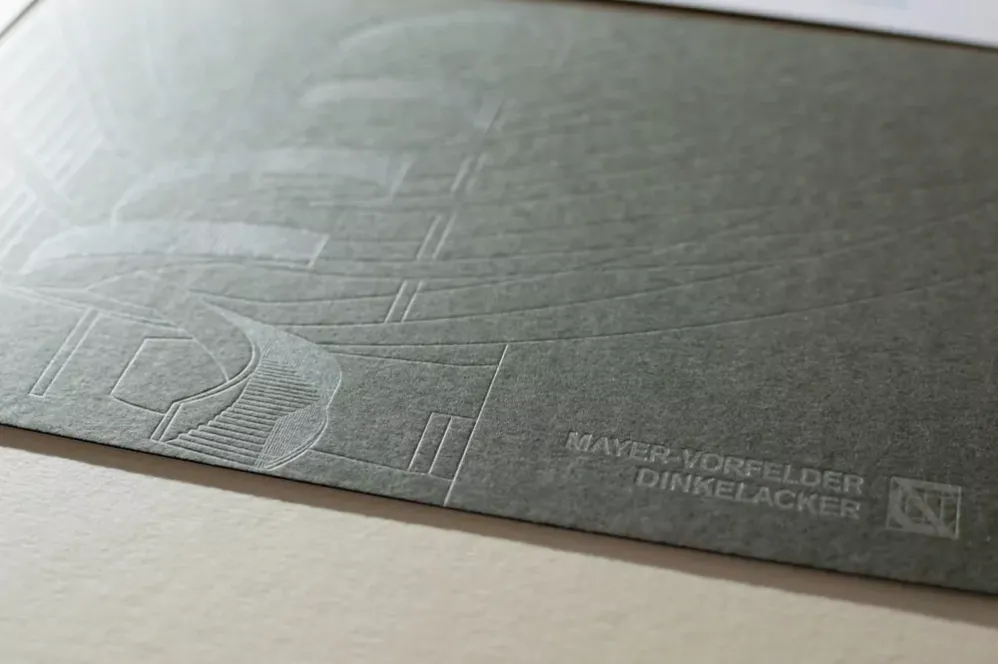 Eine grüngraue Karte mit blind geprägter Architekturzeichnung und Schriftzug im Streiflicht.