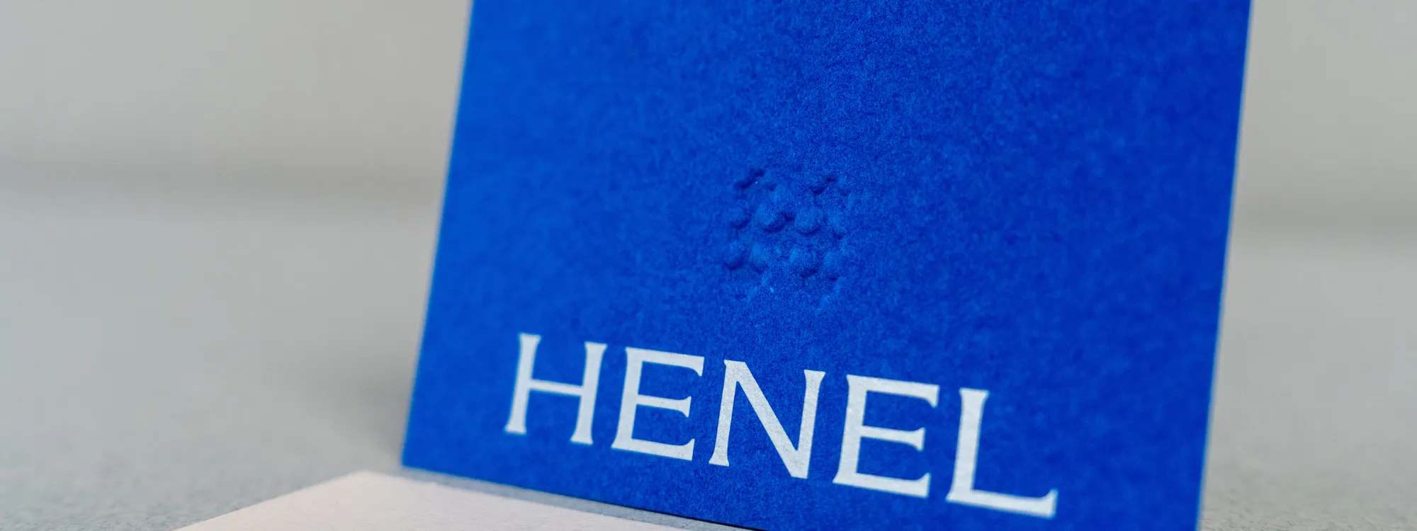 Eine royalblaue Visitenkarte aus Naturkarton mit weißem Schriftzug „HENEL“ und einem blindgeprägten Punktmuster.