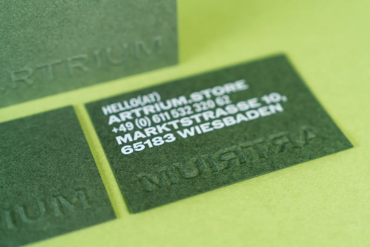 Dunkelgrüne Visitenkarten mit feiner weißer Adresszeile und blindgeprägtem Schriftzug „Artrium“ auf hellgrünem Grund.