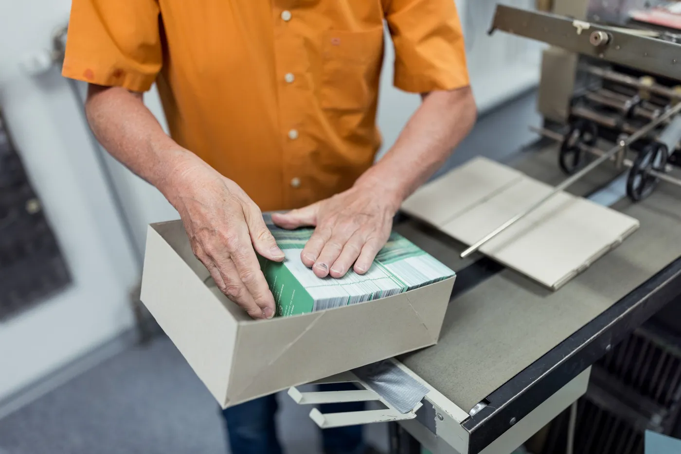 An der Auslage der Falzmaschine setzt ein Mitarbeiter einen Stapel gefalzter grüner Folder in einen Karton.