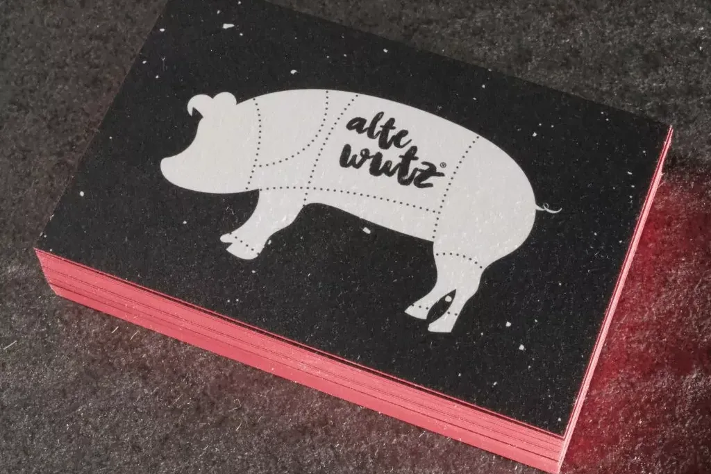 Ein Stapel schwarzer Visitenkarten mit weißem Schweinemotiv und leuchtend roten Kanten auf dunklem Stein.