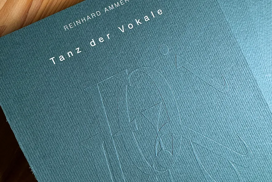 Petrolgrüne Buchumschläge „Tanz der Vokale“ mit großen, blind geprägten Buchstaben liegen auf einem Holztisch.