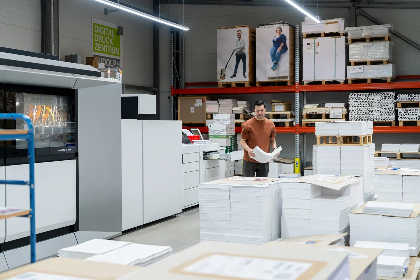 Ein Drucker blättert zwischen Stapeln frisch gedruckter Bogen neben der Inkjet-Maschine, dahinter Paletten im Hochregal.