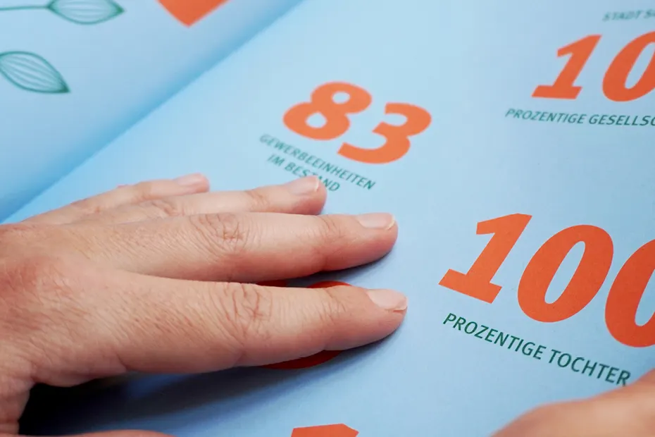 Eine Hand streicht über eine aufgeschlagene, hellblau bedruckte Broschüre mit großen orangefarbenen Zahlen.