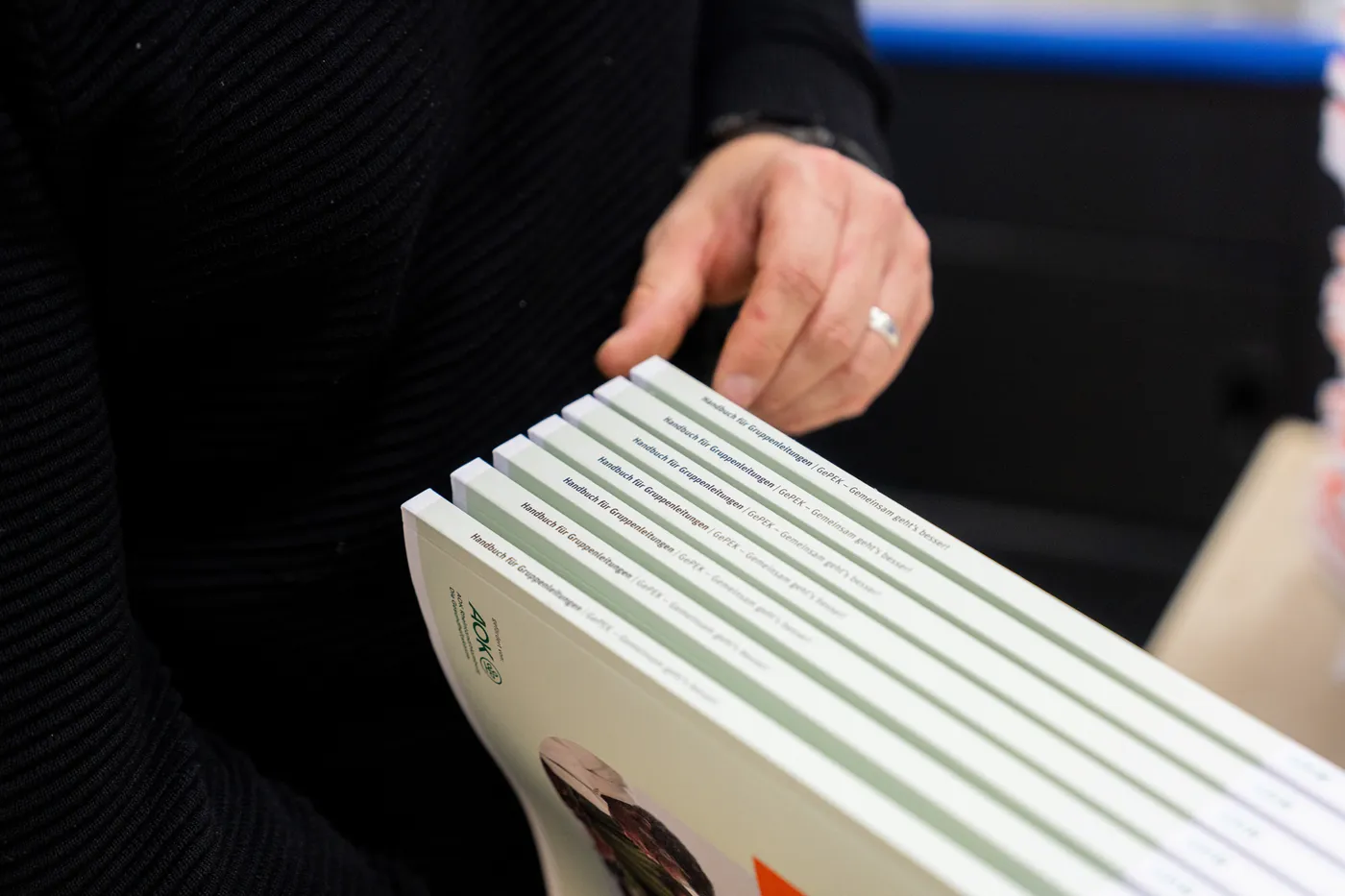 Eine Hand fächert einen Stapel klebegebundener Handbücher auf, auf jedem Rücken ist der Titel gedruckt.