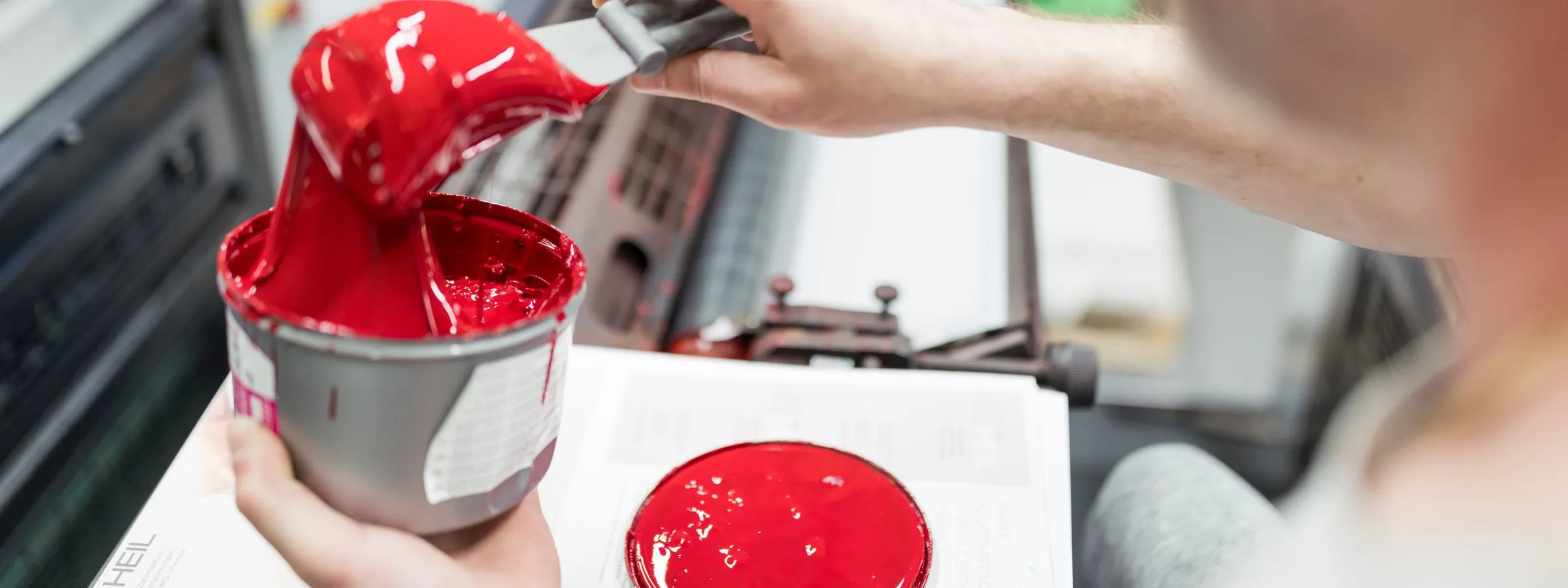 Ein Drucker nimmt mit dem Spachtel leuchtend rote Offsetfarbe aus der Dose, daneben ein Farbklecks auf dem Farbfächer.