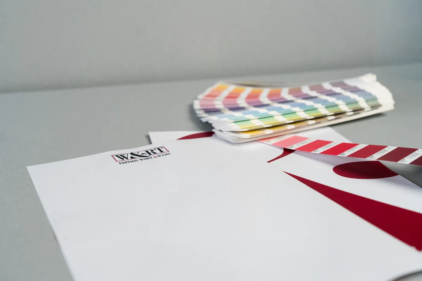 Ein Briefbogen mit weinroter Vollfläche liegt vor einem aufgefächerten Pantone-Farbfächer.