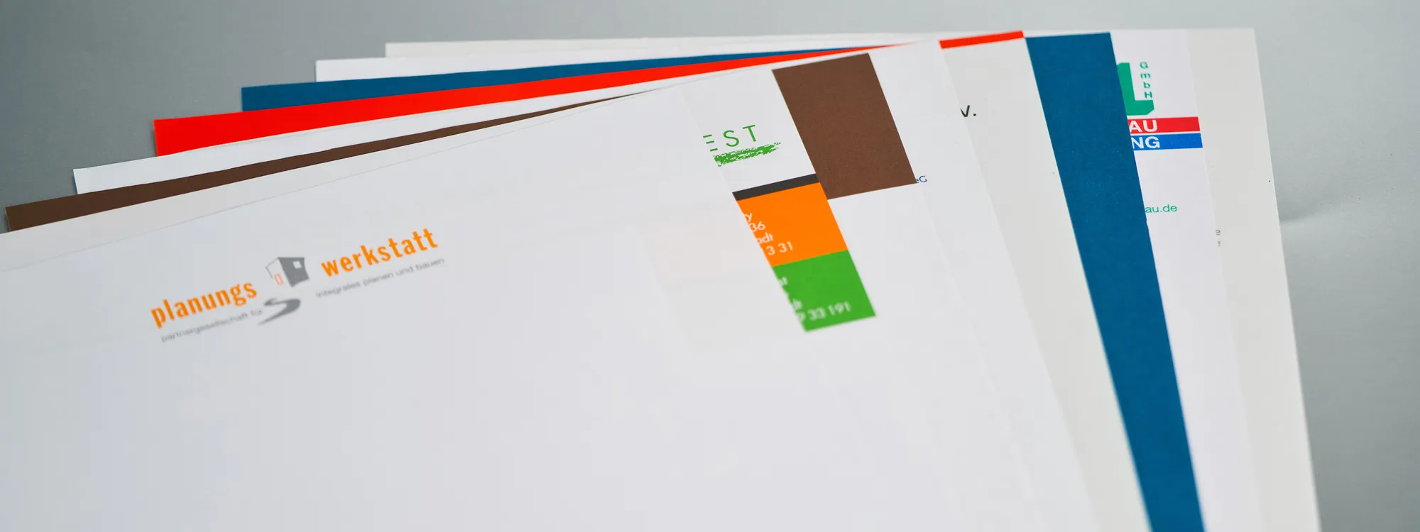 Aufgefächerte Briefbogen verschiedener Firmen, jeder mit einer eigenen Hausfarbe in Orange, Rot, Blau, Braun oder Grün.