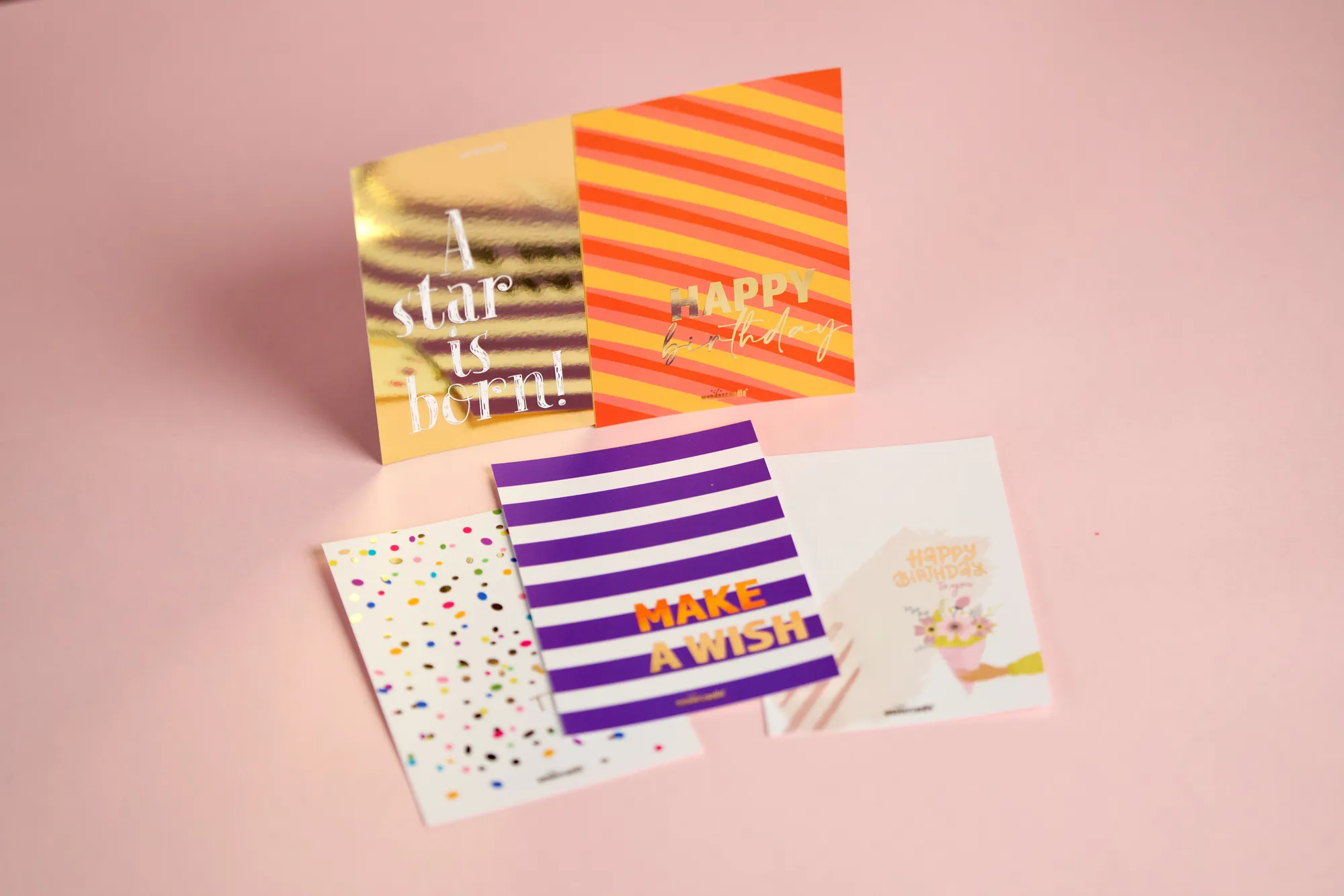 Mehrere bunt bedruckte Grußkarten mit Gold-Heißfolie liegen und stehen vor rosa Hintergrund.