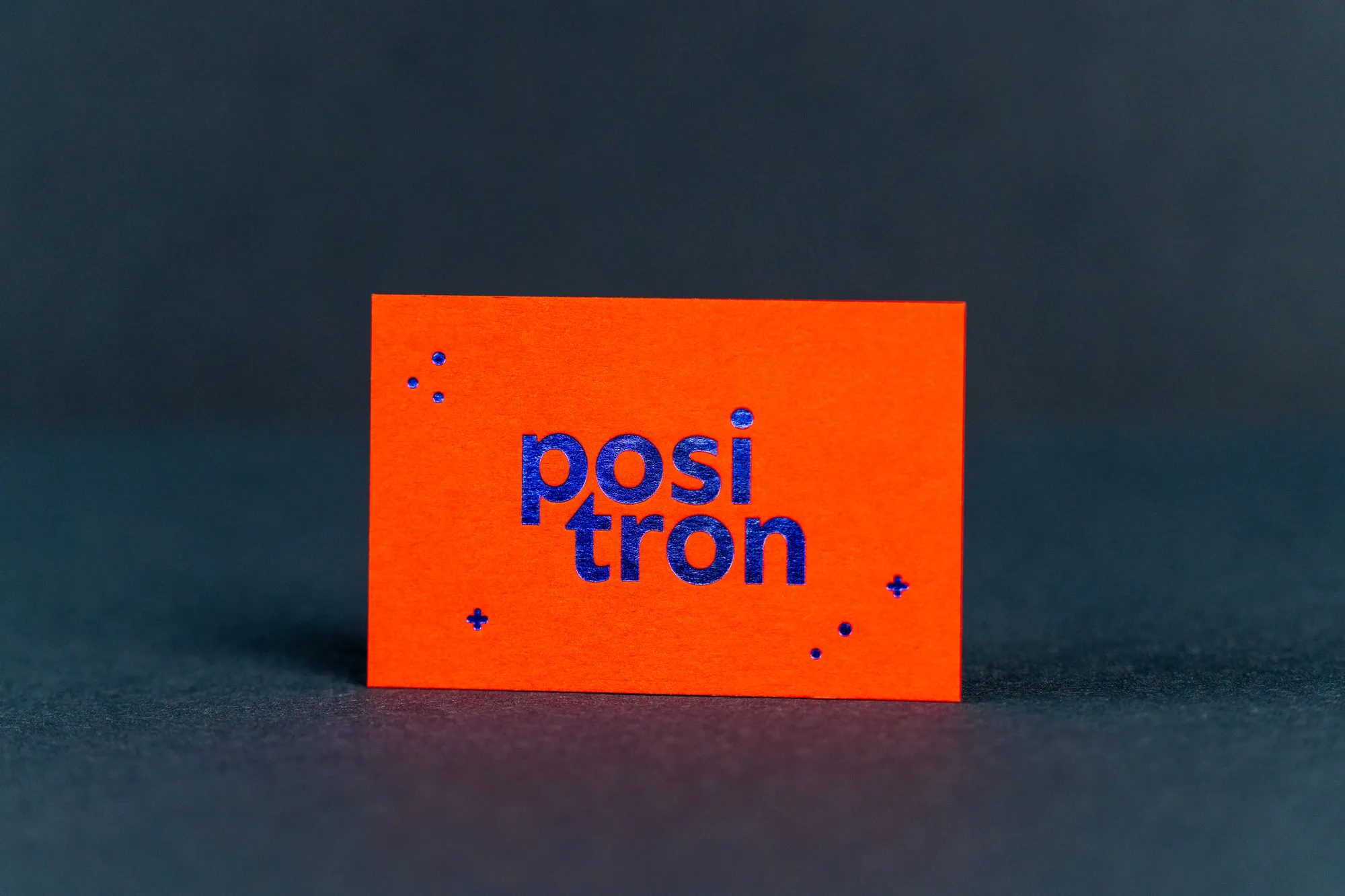 Leuchtend rote Visitenkarte mit dem Schriftzug „positron“ in blauer Heißfolie vor dunklem Grund.