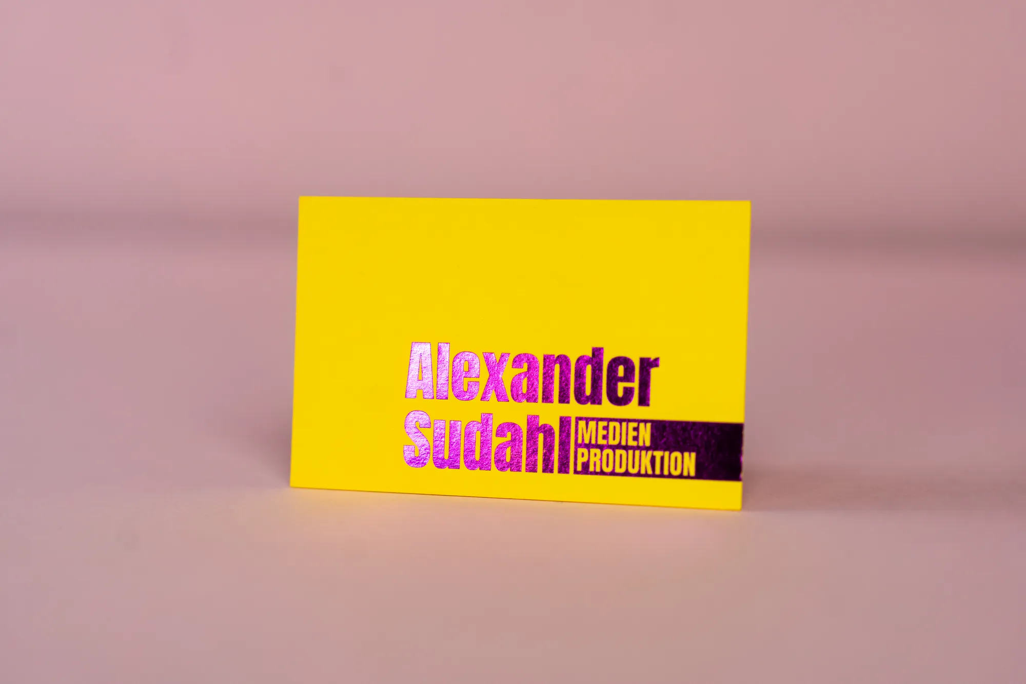 Gelbe Visitenkarte mit magentafarbener Heißfolie „Alexander Sudahl“ auf rosa Grund.