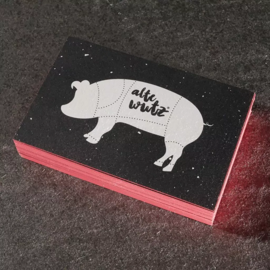 Stapel Visitenkarten mit einem Schwein als Motiv und rotem Farbschnitt auf dunklem Stein.