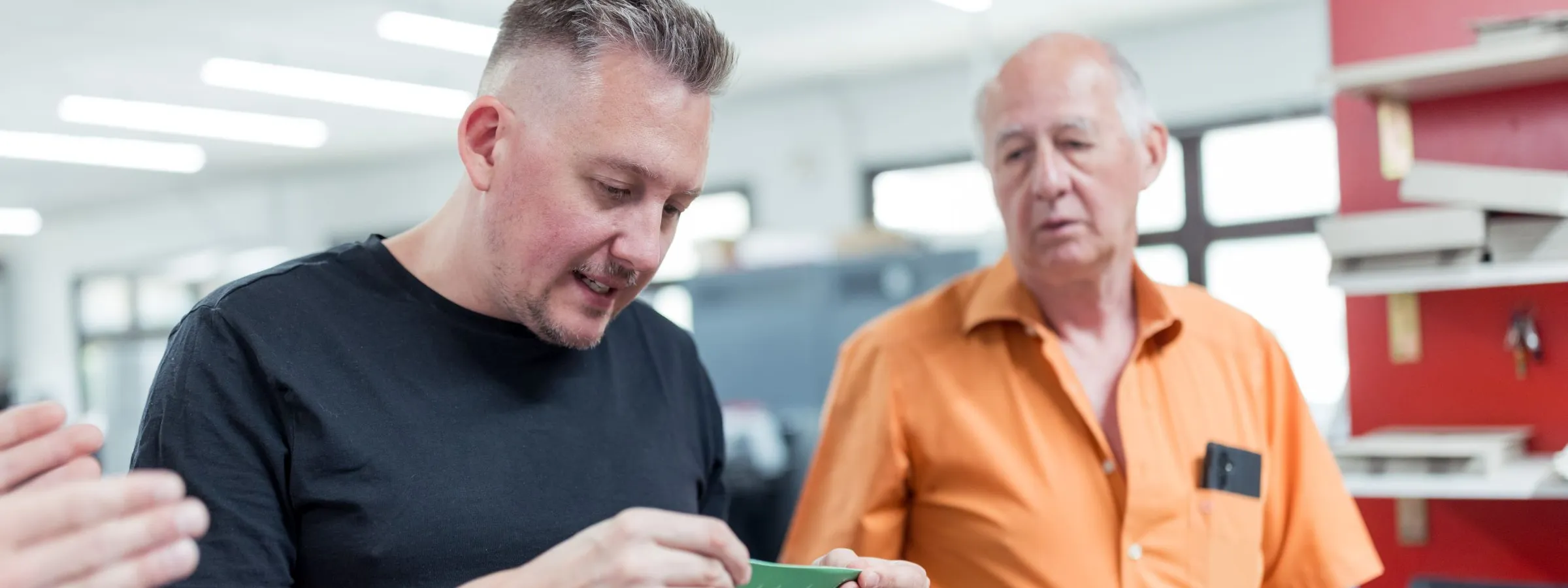 Ein Mitarbeiter prüft einen grünen, gerillten Folder in den Händen, ein älterer Kollege im orangefarbenen Hemd schaut zu.