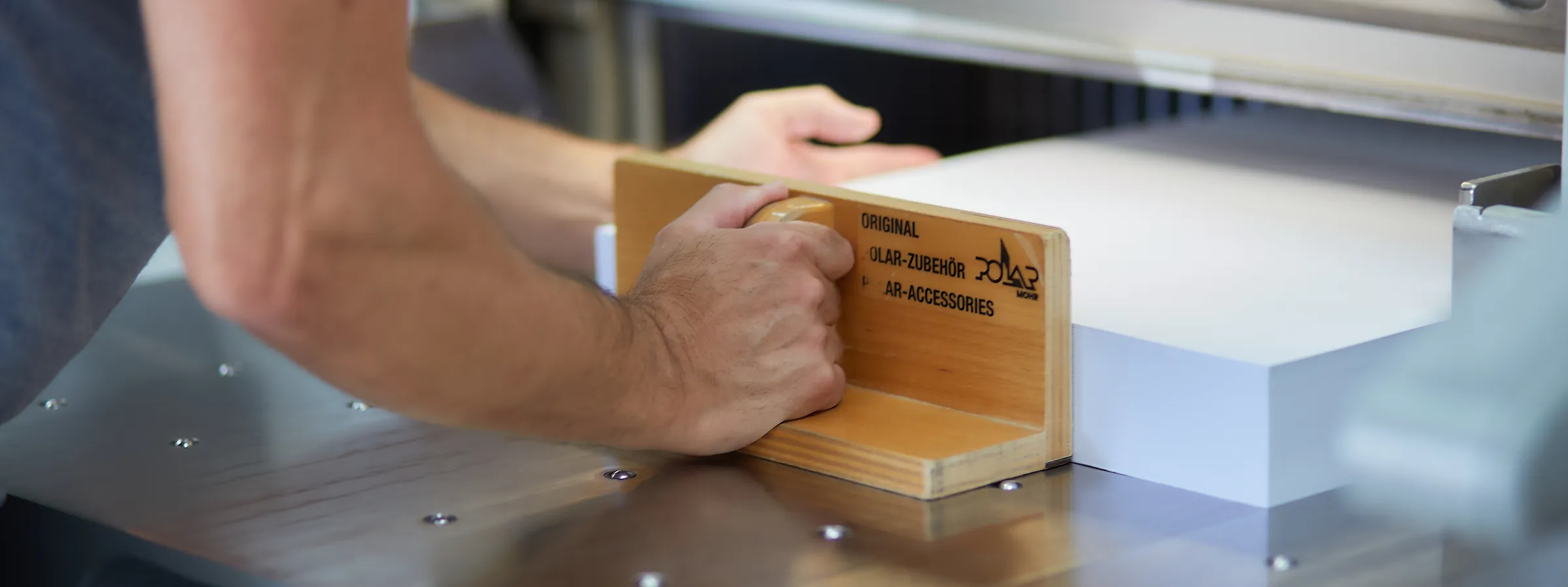 Eine Hand schiebt mit einem hölzernen Anlegewinkel einen weißen Papierstapel unter den Pressbalken des Planschneiders.