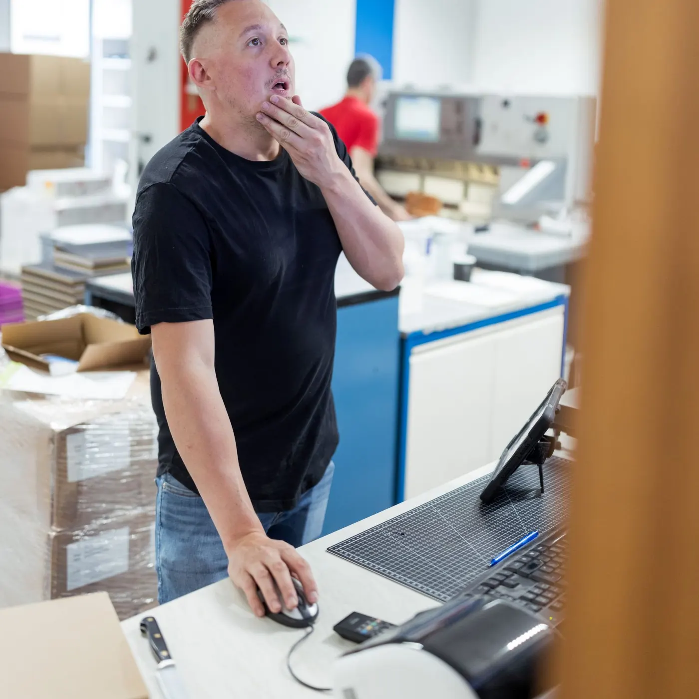 Ein Drucker steht nachdenklich an seinem Arbeitsplatz mit Schneidematte und Bildschirm, im Hintergrund die Schneidemaschine.