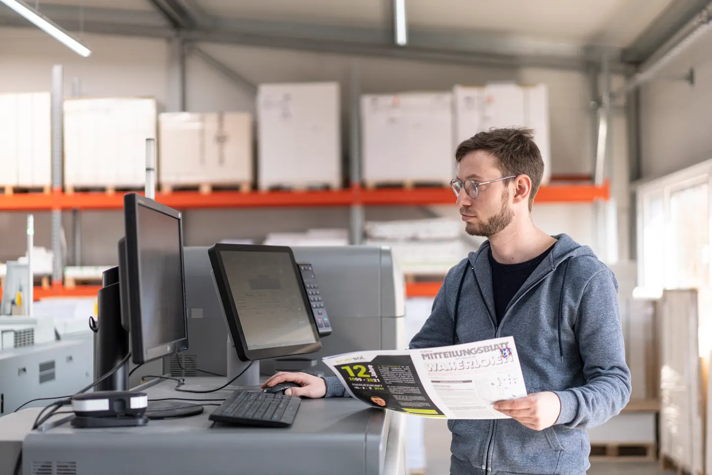 Ein Drucker hält einen gedruckten Bogen in der Hand und vergleicht ihn mit dem Bildschirm am Leitstand.