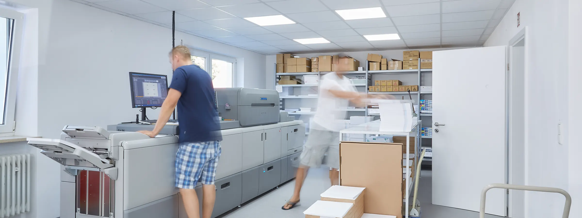 Ein Drucker steht an der Bedienkonsole einer Toner-Digitaldruckmaschine, im Hintergrund trägt ein Kollege frisch gedruckte Bogen zum Regal.