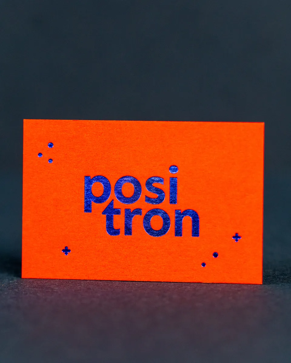 Eine leuchtend orange Visitenkarte mit dem Schriftzug „positron“ in blauer Heißfolie steht vor dunklem Hintergrund.