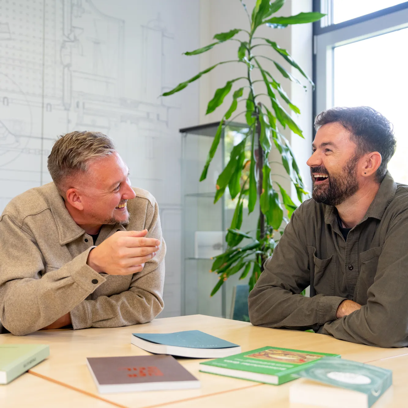 Zwei Kollegen lachen am Besprechungstisch, vor ihnen liegen gedruckte Bücher.