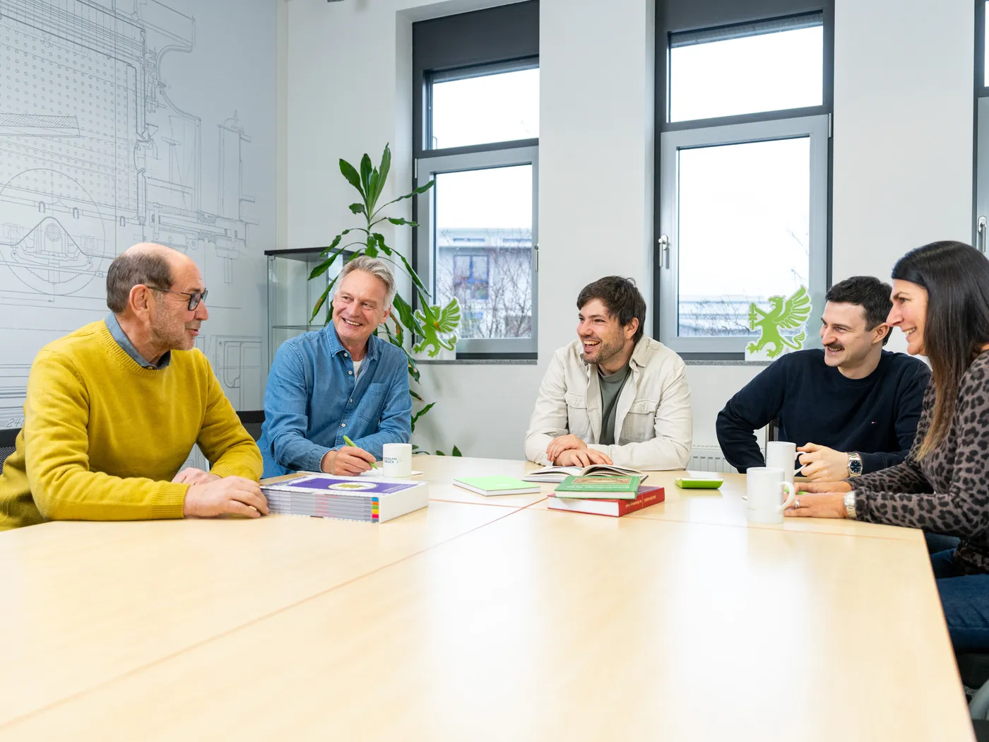 Fünf Kolleginnen und Kollegen sitzen lachend am Besprechungstisch, vor ihnen liegen gedruckte Bücher.