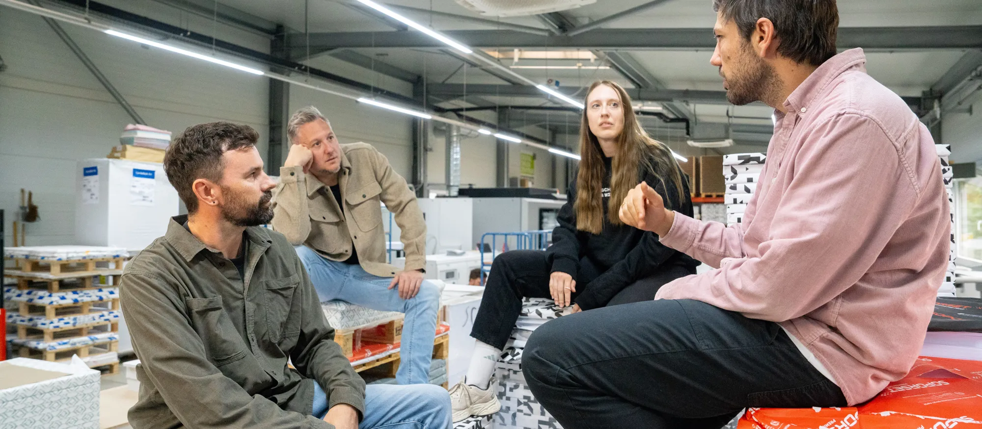 Vier Kollegen sitzen im Papierlager auf Paletten mit Papierpaketen und sprechen miteinander.